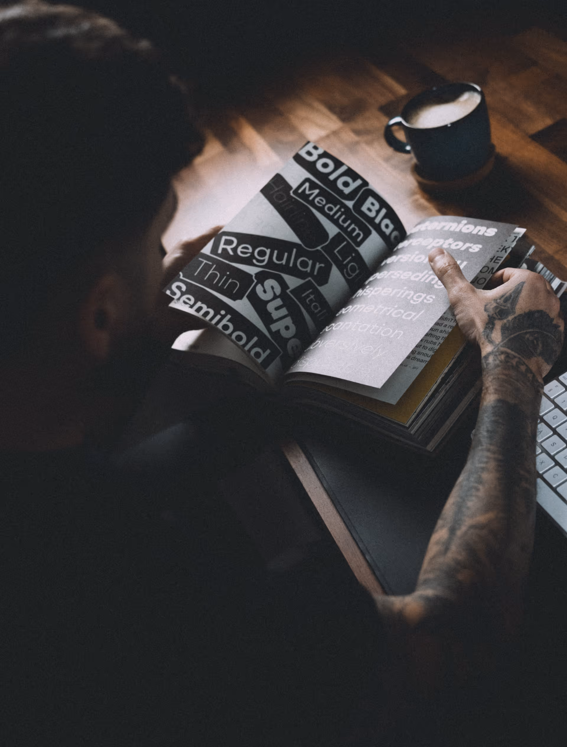 Tattooed man reading a typography book with words like Bold, Black, Medium, Regular, Thin, and Semibold, next to a cup of coffee on a wooden table.